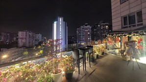 Une terrasse sur le toit, agrémentée de plantes en pot, d’une table et de chaises, d’où l’on peut admirer la vue sur la ville illuminée la nuit.