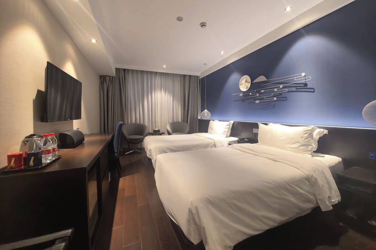 Een hotelkamer met twee bedden, een bureau met stoel, een televisie en een ventilator aan de muur.
