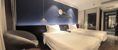 A modern hotel room with two beds, a dark blue accent wall, a round pendant light, and a small table with a lamp.