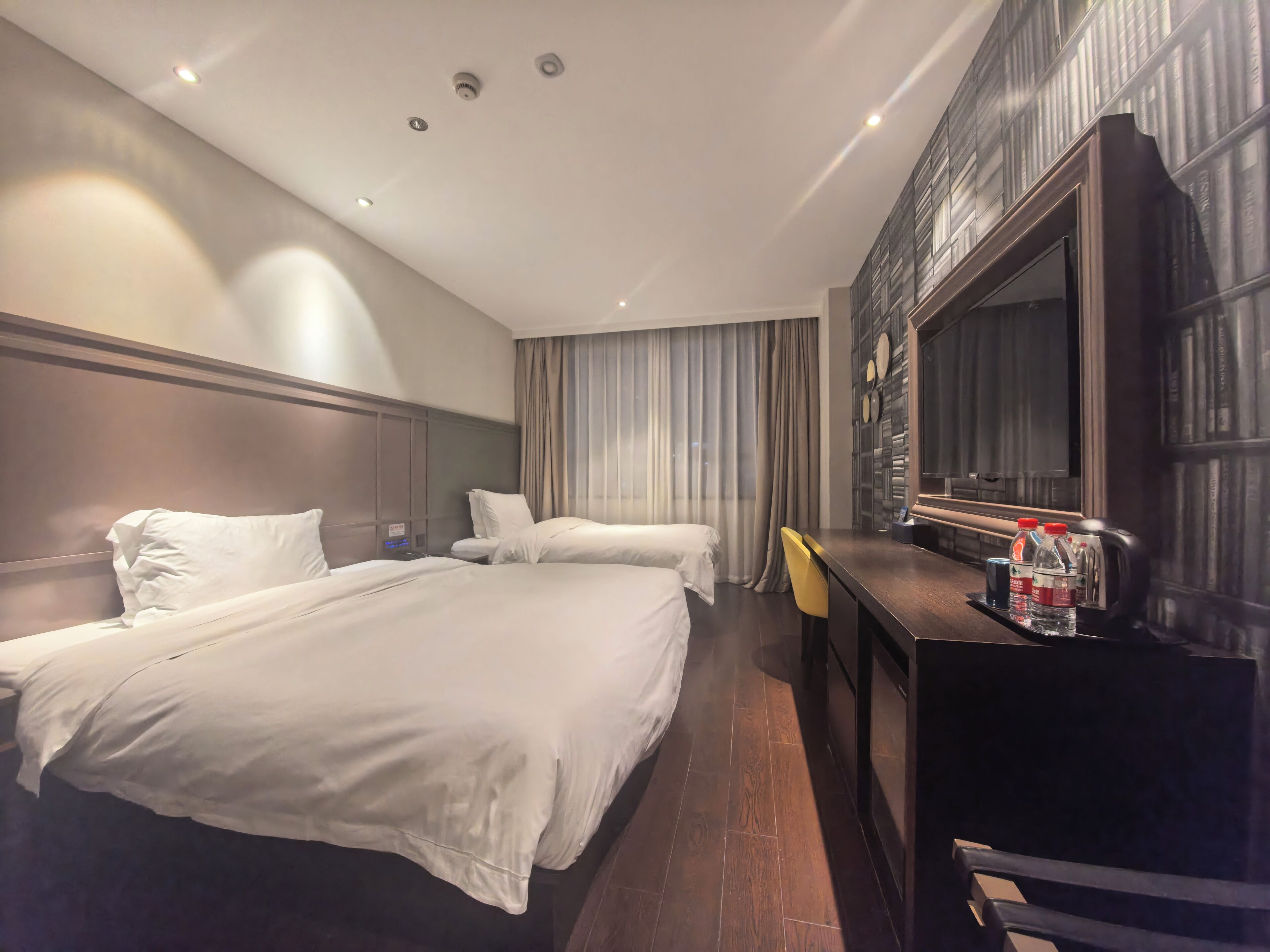 A hotel room with two beds, a desk, a chair, a large mirror, and a bookshelf.