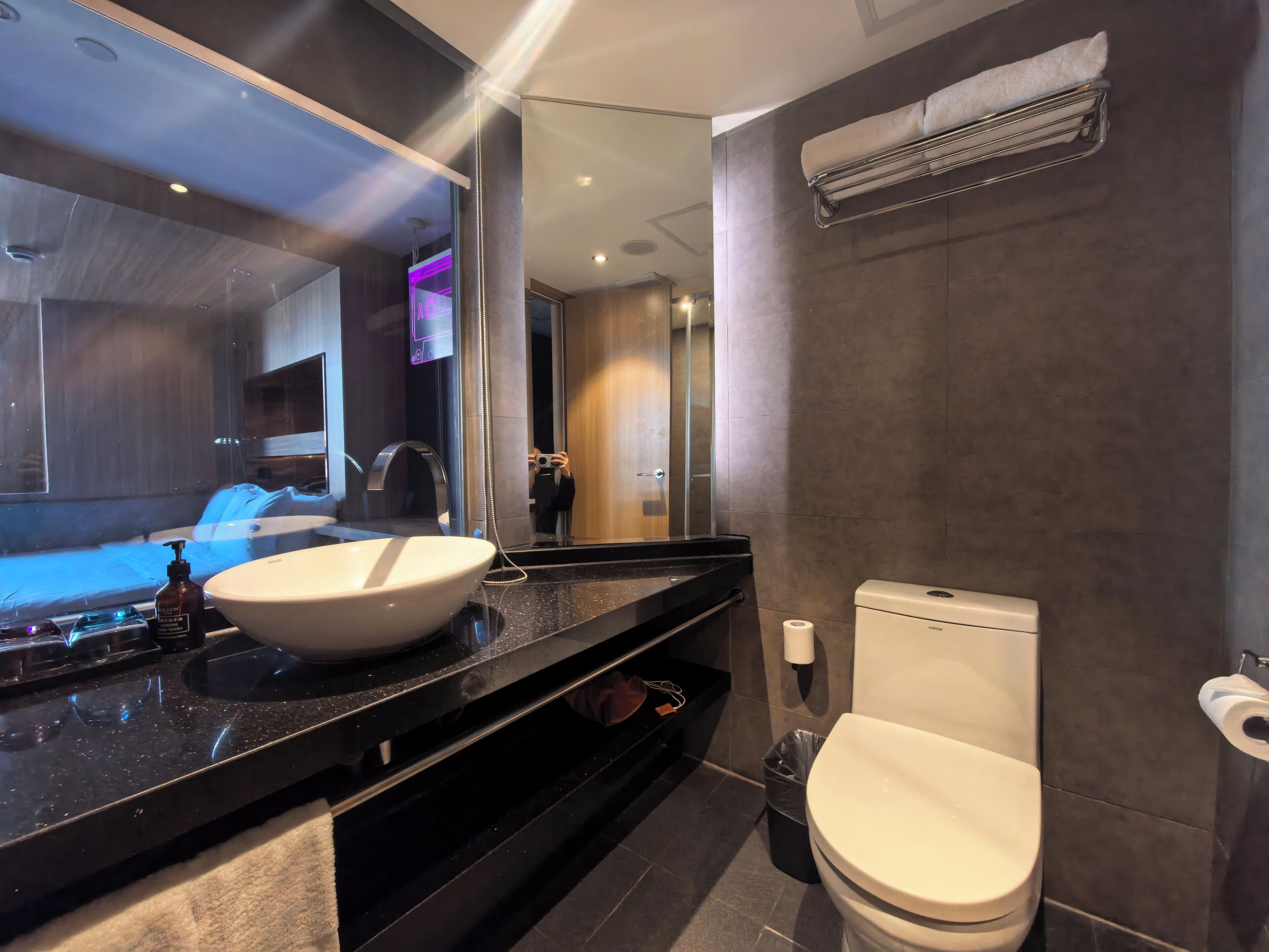 A modern bathroom with a large mirror, a white sink, a black countertop, a toilet, and a trash bin.