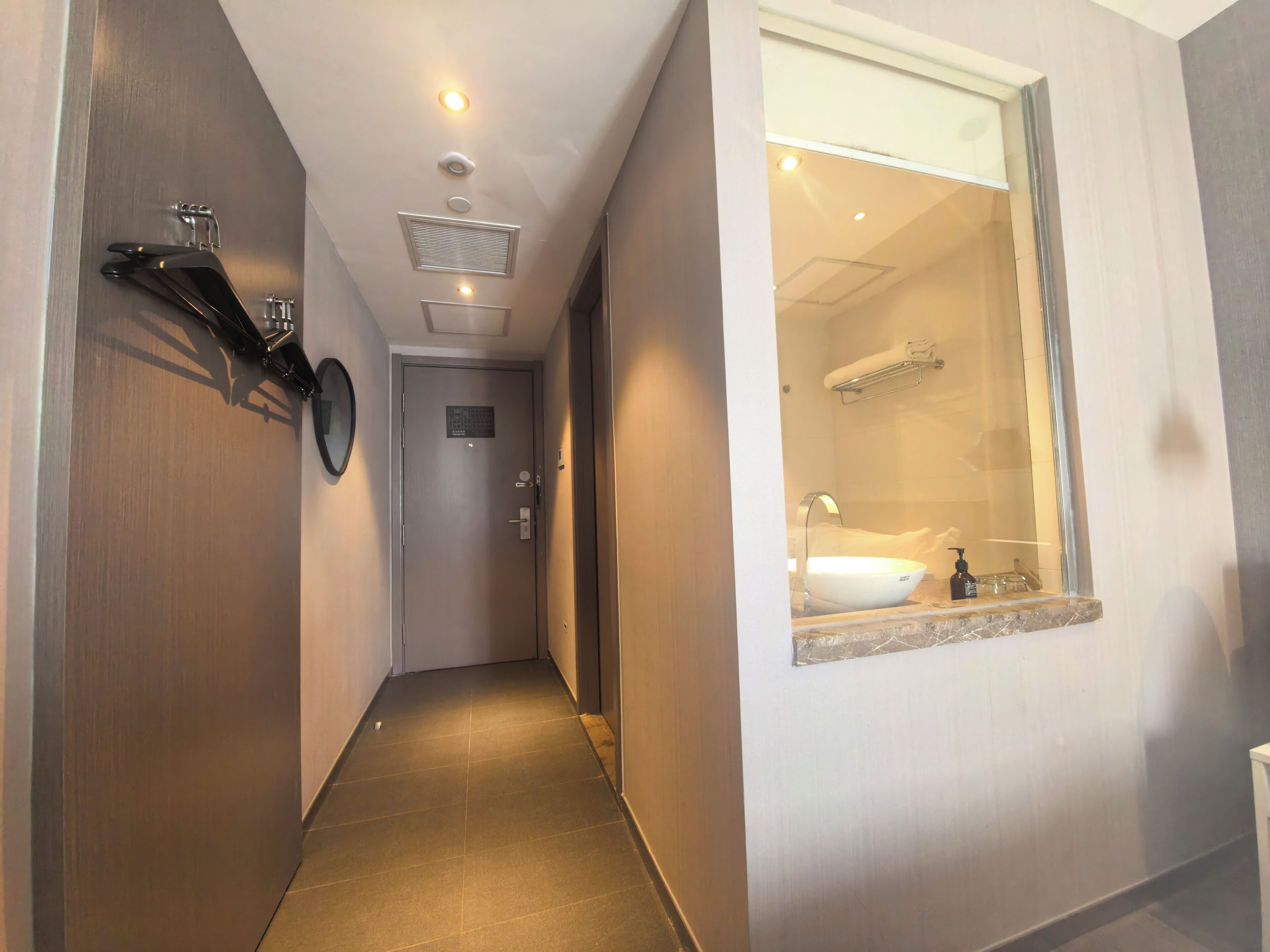 Ein schmaler Flur mit einer Tür am Ende, einem Spiegel an der linken Wand und einem Badezimmer mit Waschbecken und Handtuchhalter, das durch ein Fenster sichtbar ist.