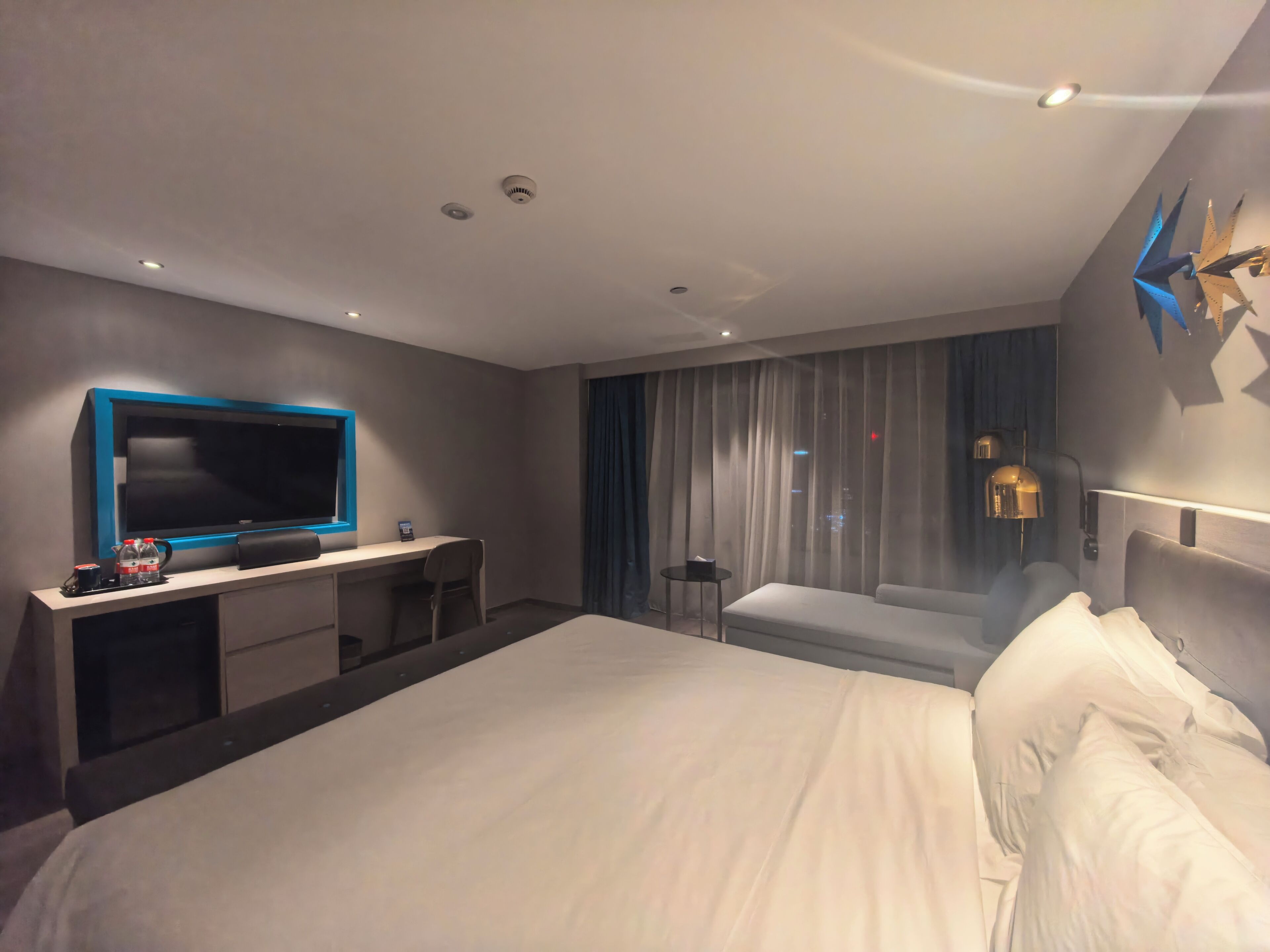 A modern hotel room with a large bed, a desk with a flat-screen TV, a small table, and a large window with curtains.