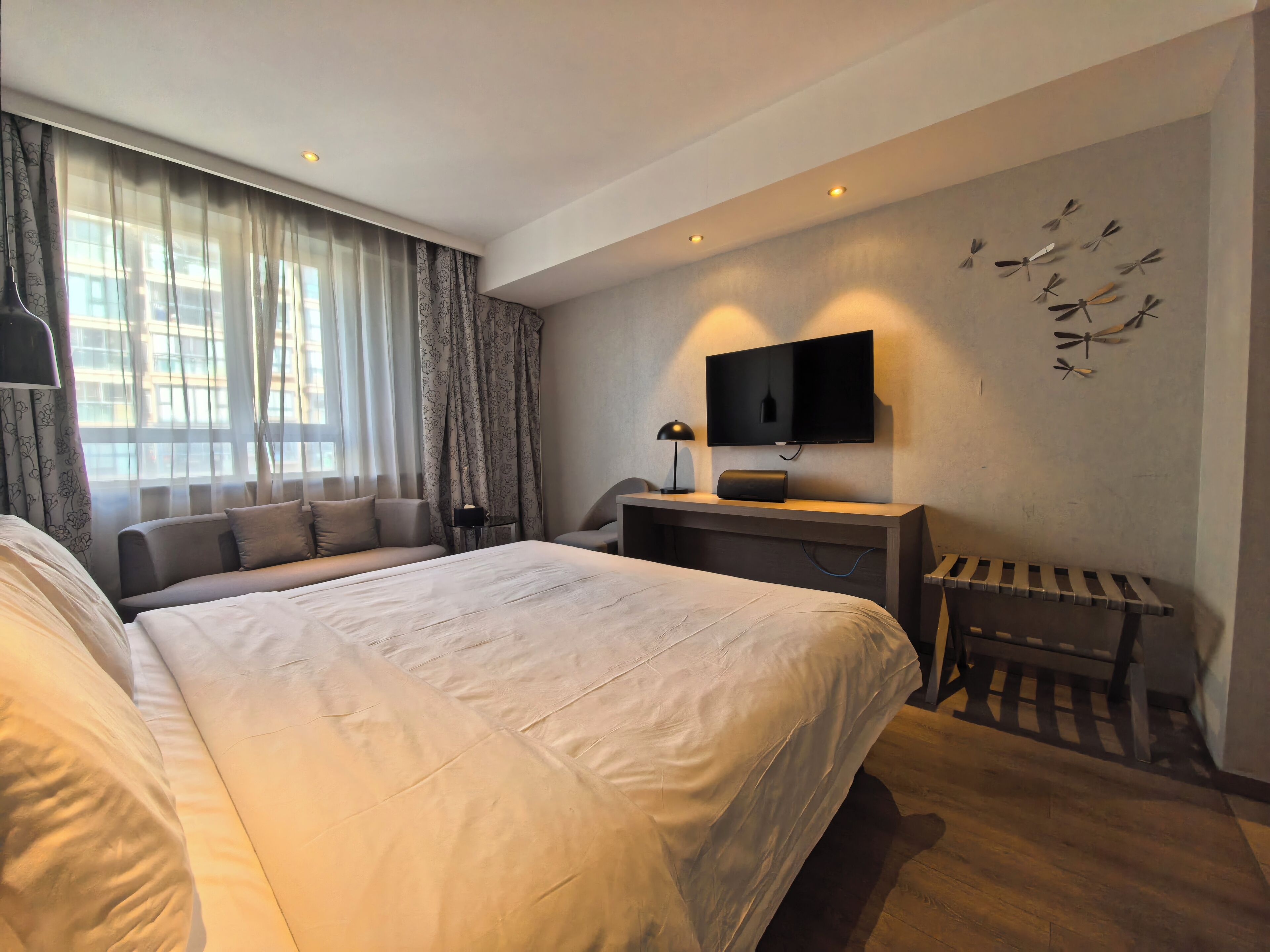 A modern hotel room with a large bed, a sofa, a desk with a flat-screen TV, and a decorative wall piece.