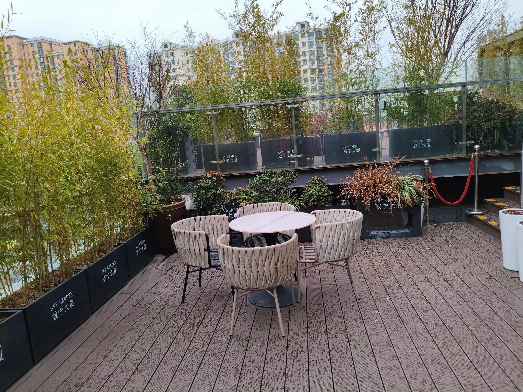 A rooftop terrace with a round table and chairs, surrounded by potted plants and a glass railing.