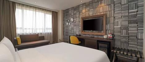 A hotel room with a large bed, a TV, a desk with a chair, and a sofa.