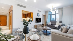 A modern living room with a glass dining table set for four, a sofa, a TV, and a city view through large windows. - Modern Retreat in The Views with Balcony & Pool (Dubai)