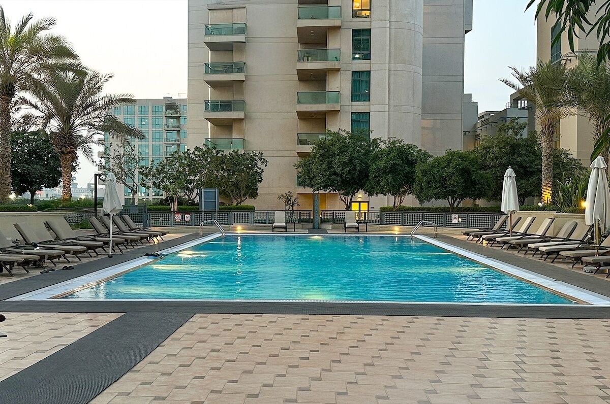Una piscina rectangular amb gandules i ombrel·les, envoltada d'arbres i un edifici alt al fons.