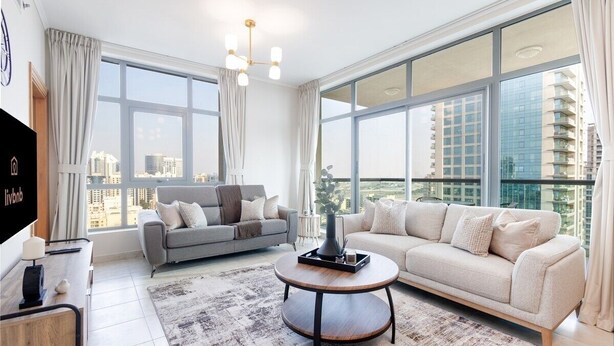 Una sala de estar moderna con un sofá, una mesa de centro y un televisor. Amplias ventanas ofrecen vistas de la ciudad.