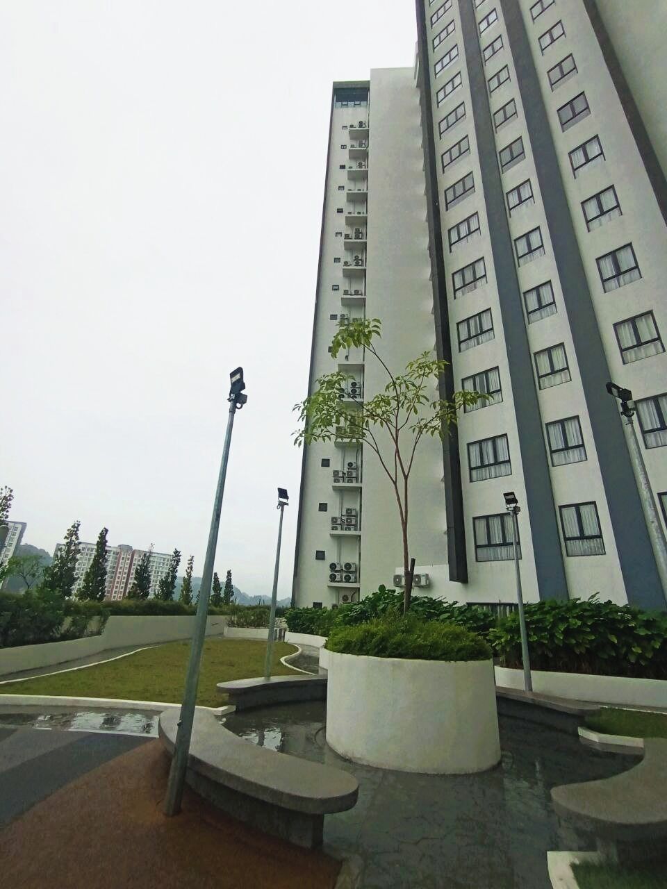 A high-rise residential building with multiple balconies, a landscaped area with a small tree, and a street lamp.