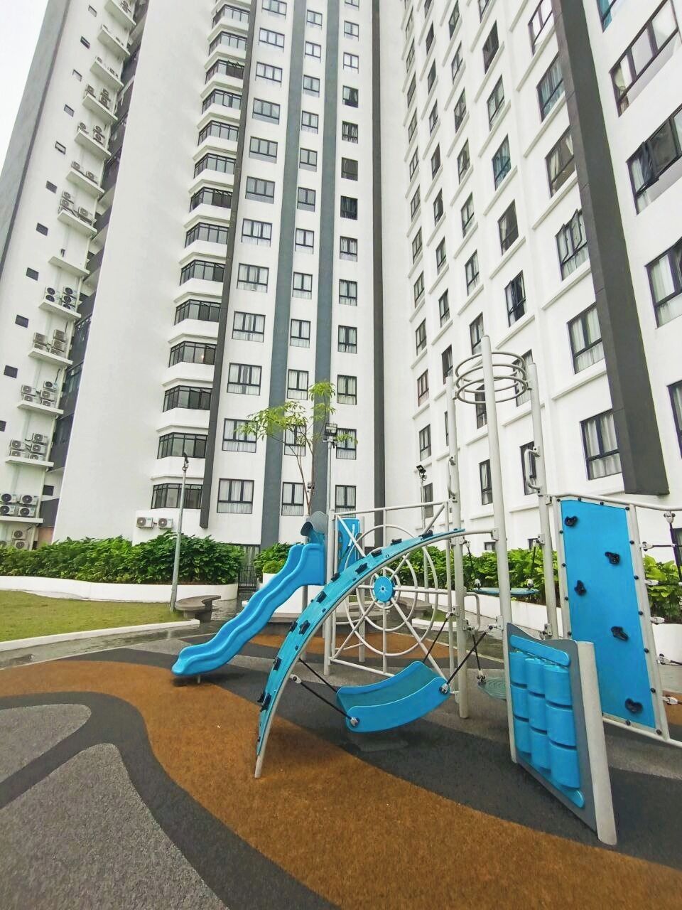 A playground with a slide and climbing structures, surrounded by a landscaped area with a tall residential building in the background.