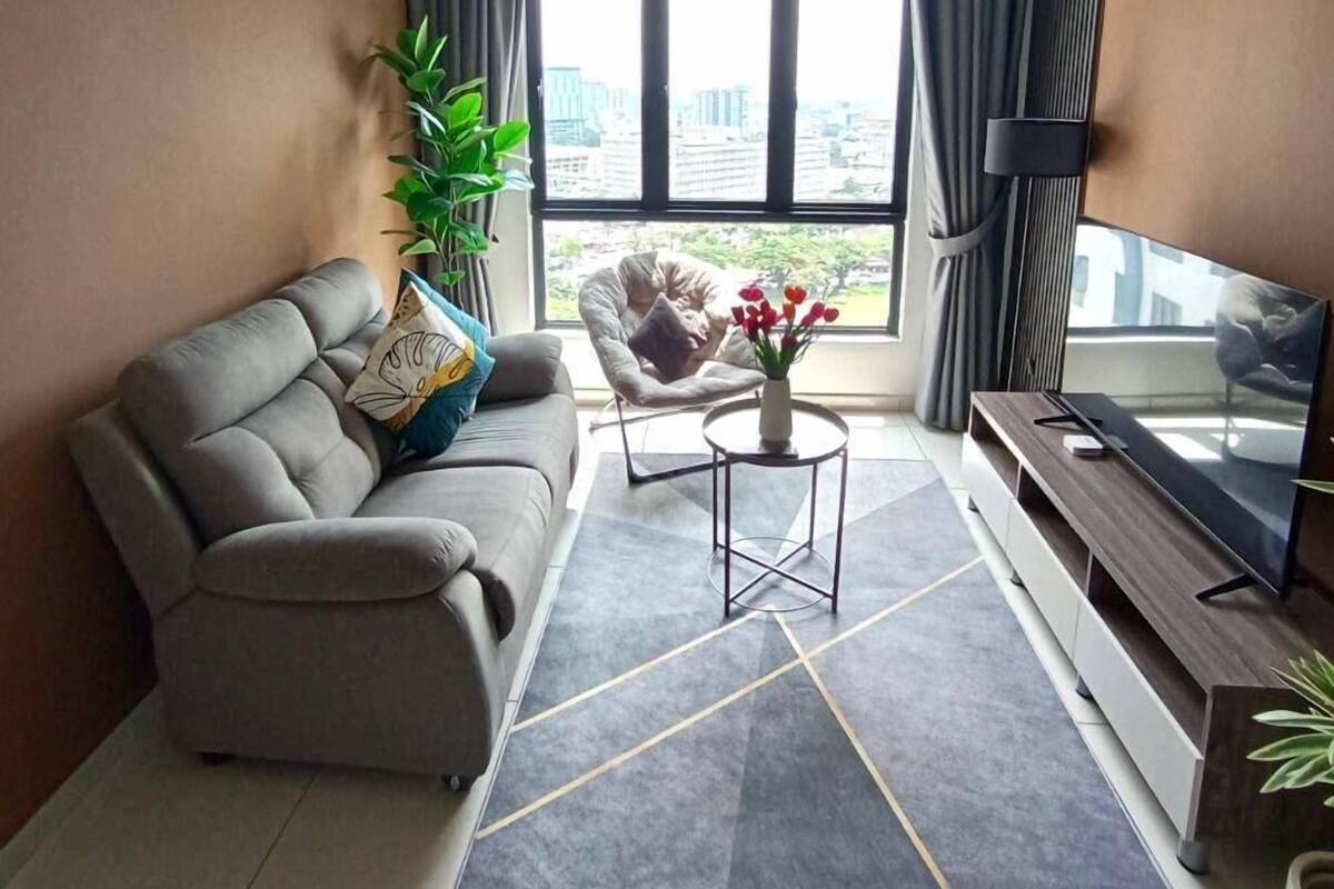 Een moderne woonkamer met een grijze bank, een klein bijzettafeltje, een televisie op een houten dressoir, een plafondventilator en een groot raam met gordijnen.