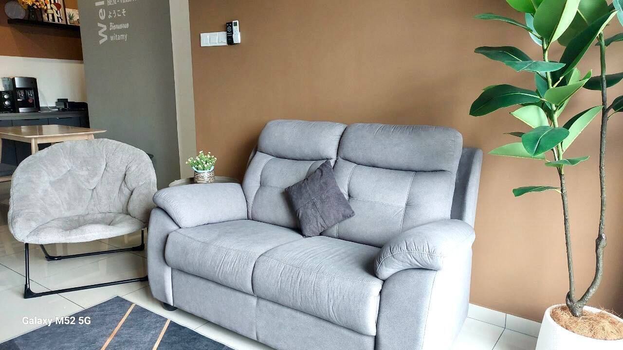 A grey sofa with a cushion, a grey armchair, a small table, and a potted plant in a living room with a brown wall and tiled floor.