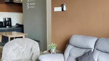 A modern living room with a grey sofa, a white armchair, and a small side table with a plant. A coffee machine is visible in the background.