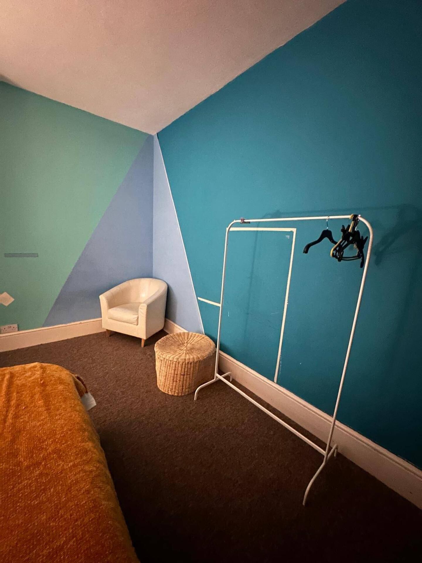 Quarto com uma cama, uma cadeira, um pequeno banquinho e uma estante de metal em frente a uma parede azul.