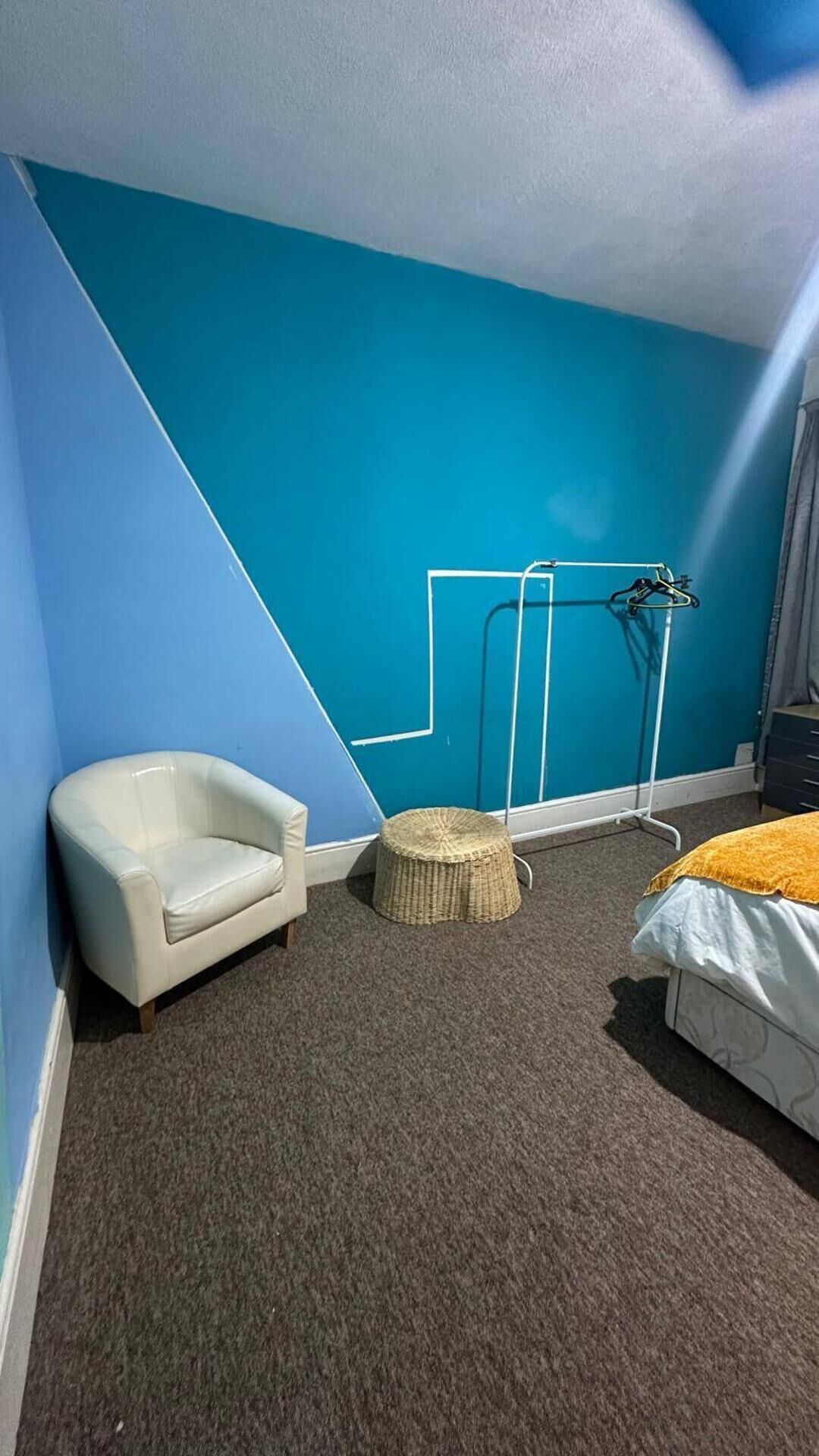Una habitación con una pared azul, un sillón blanco, un taburete redondo de mimbre y una cama con una manta amarilla.