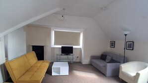 A small, sparsely furnished living room with a yellow sofa, a grey sofa, a white coffee table, and a white armchair. There is a television on a glass table, a window with blinds, and a ceiling light. - Southampton Serenity - 3 Bedroom Apartment (Southampton)