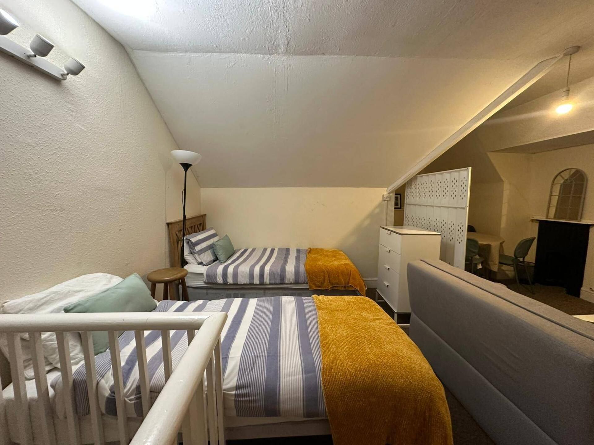 A small, triangular attic bedroom with a crib, two beds with striped bedding, a floor lamp, and a mirror reflecting a dining area.