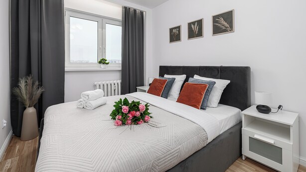 A modern bedroom with a large bed, grey curtains, a bedside table with a lamp, and three framed pictures on the wall. - Lumina 3-room apartment with balcony (Warsaw)