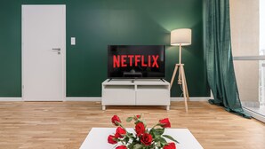 A modern living room with a flat-screen TV displaying 'NETFLIX', a white entertainment unit, a floor lamp, and a vase of red roses on a white table. - Lumina 3-room apartment with balcony (Warsaw)