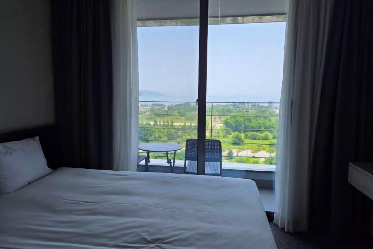 A hotel room with a large bed, a view of a balcony with a table and chairs, and a cityscape in the background.