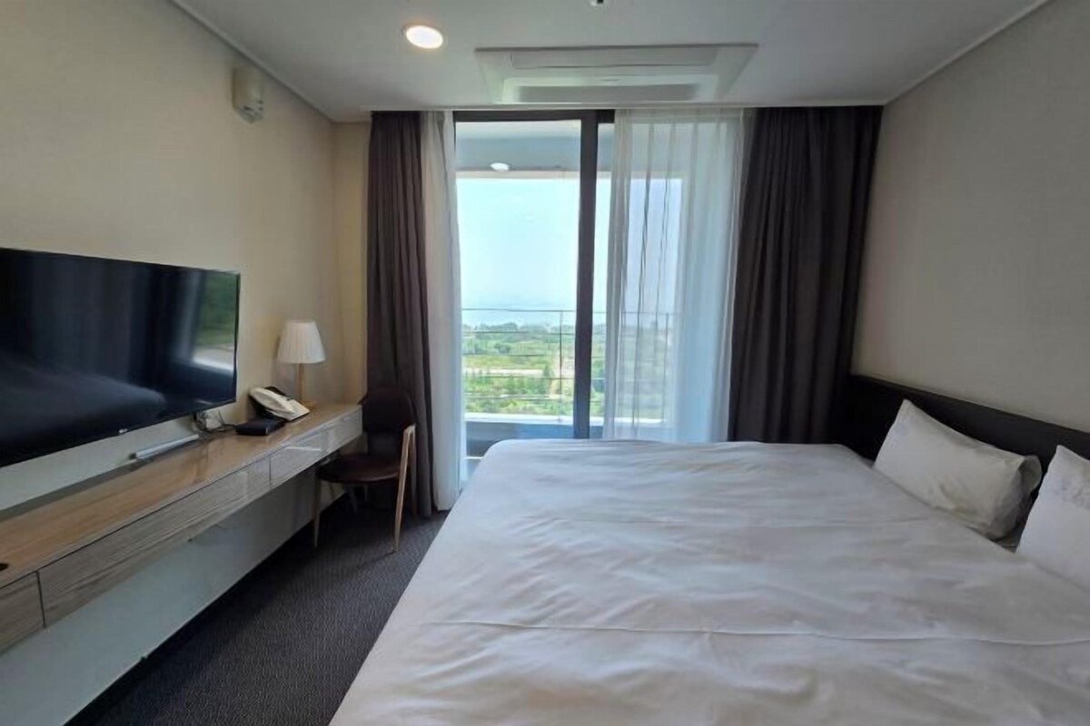 Un cuarto de hotel con una cama grande, un televisor sobre un aparador de madera, un escritorio con lámpara y vistas al exterior a través de un ventanal.