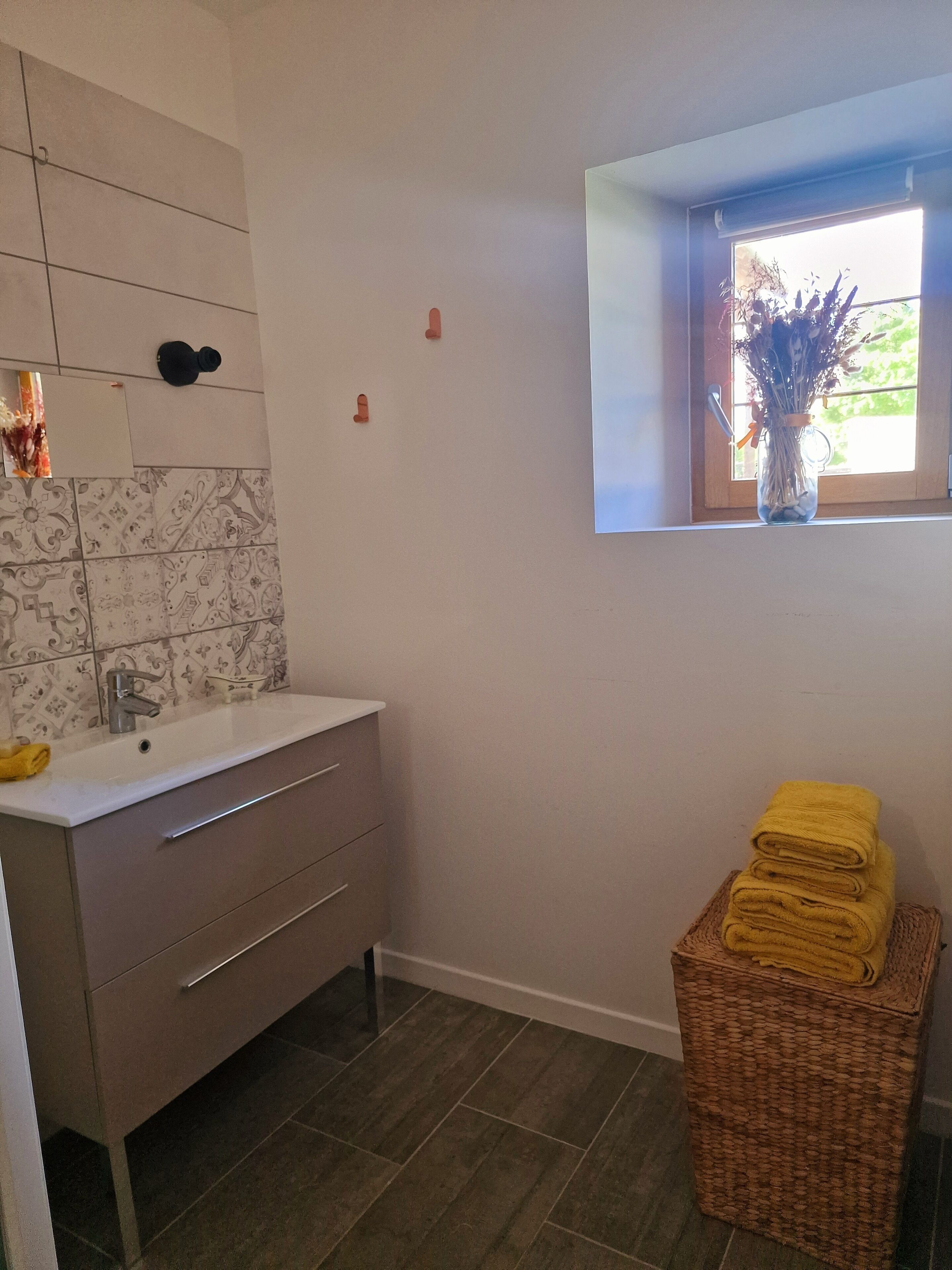A bathroom with a white sink, a mirror, a window with a view of greenery, and a wicker basket with yellow towels.