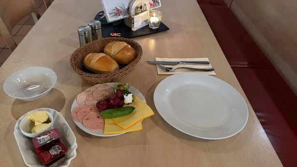 A table set with a plate of sliced meats, cheese, grapes, and a cucumber, along with condiments and a basket of bread. - Waldherberge Sargenroth  (Sargenroth)