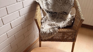 A wicker chair with a fluffy grey cushion and a wooden table in a room with a white brick wall and wooden flooring.