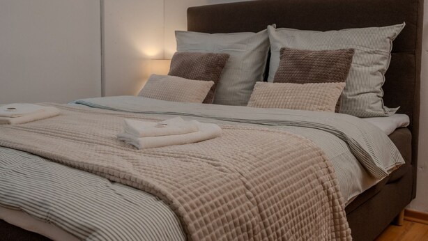 A neatly made bed with a striped blanket and pillows, a bedside table with a lamp, and a plain wall.