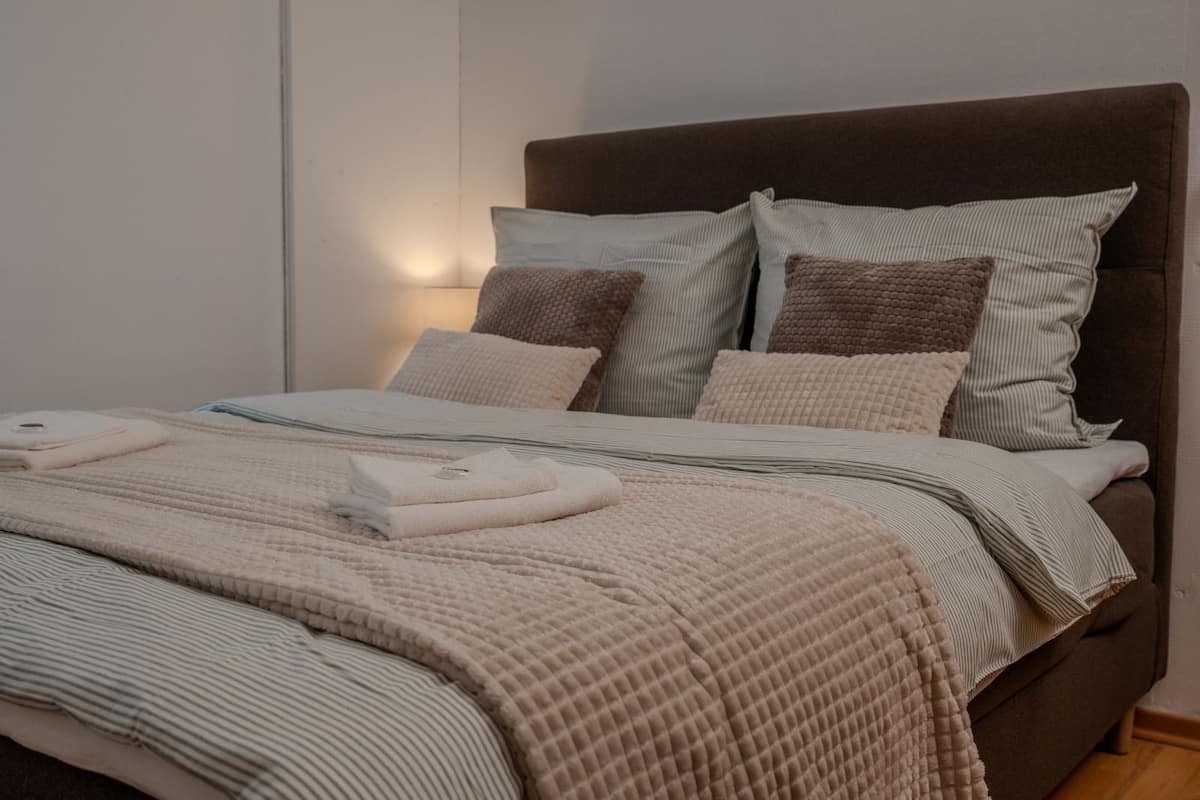 Ein ordentlich bezogenes Bett mit gestreiftem Bettbezug und Kissen, ein Nachttisch mit Lampe und eine schlichte Wand.