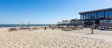 Ein Strand mit Holzbanken, einem Rettungsschwimmturm und einem modernen Gebäude mit großen Fenstern.