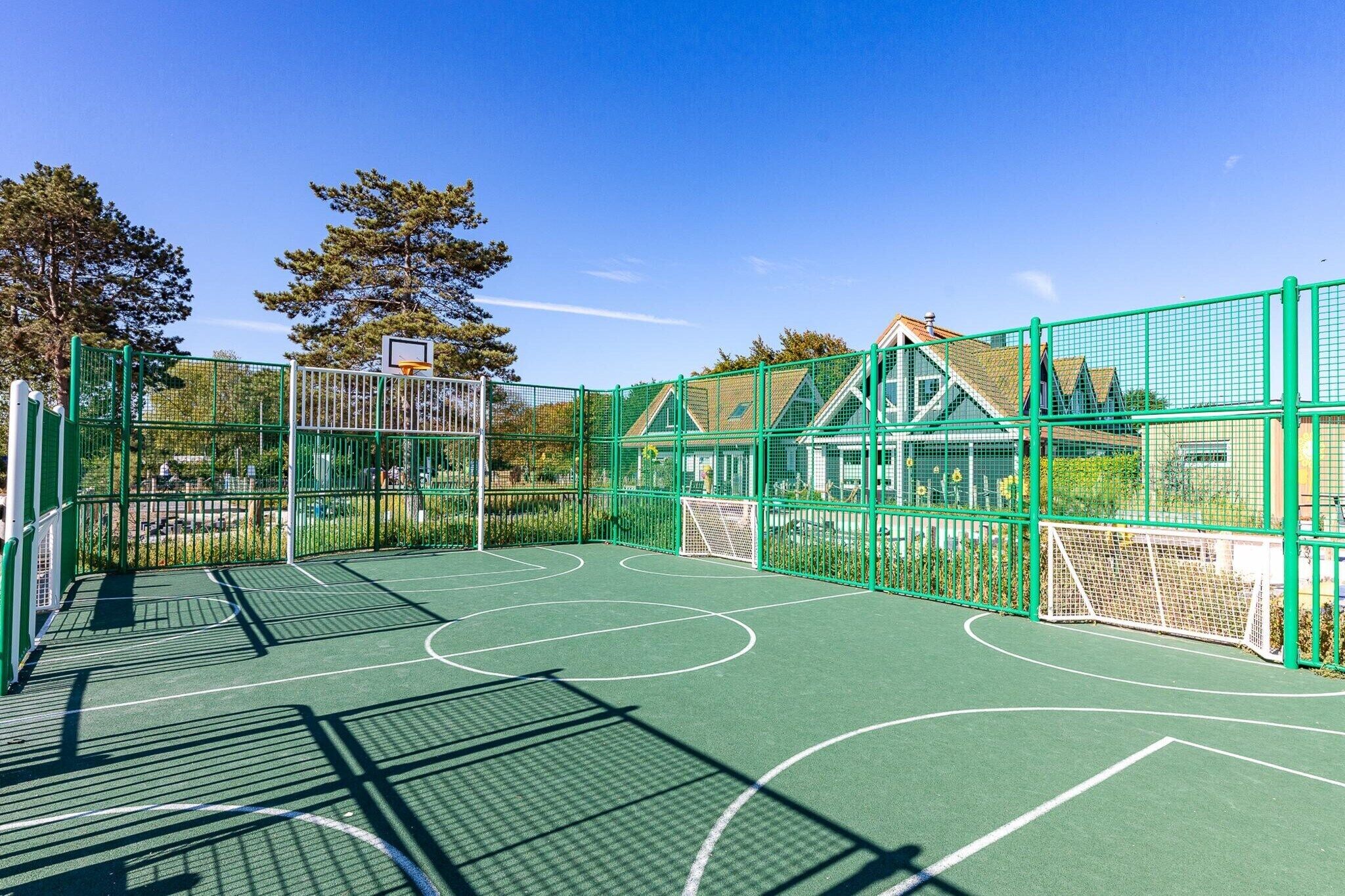 An outdoor basketball court with a green fence, a basketball hoop, and a house in the background.
