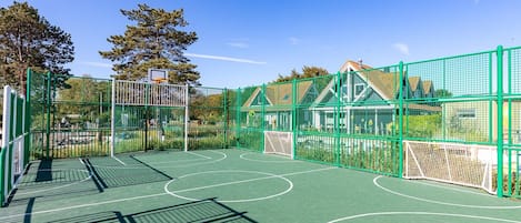 An outdoor basketball court with a green fence, a basketball hoop, and a house in the background.