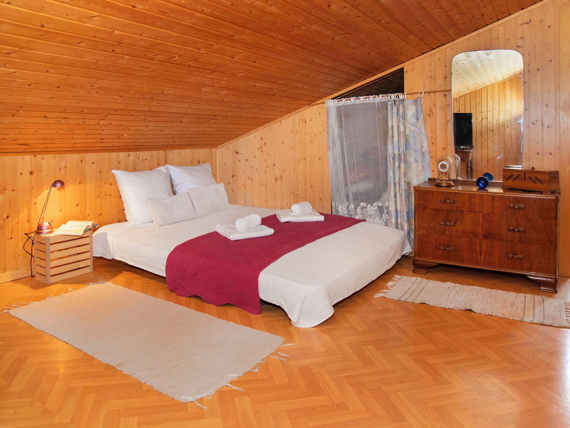 A bedroom with a wooden ceiling, a bed with white bedding and a red blanket, a wooden dresser, and a mirror.