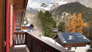Un balcone in legno con vista su un paesaggio montano, con cime innevate e alberi dai colori autunnali.