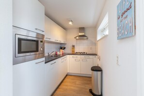A compact kitchen with white cabinets, a stainless steel microwave, a gas stove, and a trash bin. - Beachside Modern Holiday Home for 6 (Renesse)