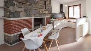 Una cucina moderna con parete in pietra, camino in mattoni, mobili bianchi e zona pranzo con tavolo in vetro e sedie bianche.