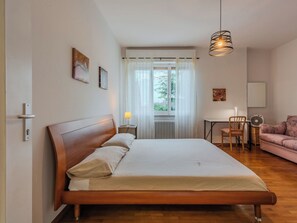 Ein Schlafzimmer mit einem hölzernen Bett, einem Nachttisch, einer Lampe, einem Sessel, einem Sofa und einem Fenster mit Vorhängen.