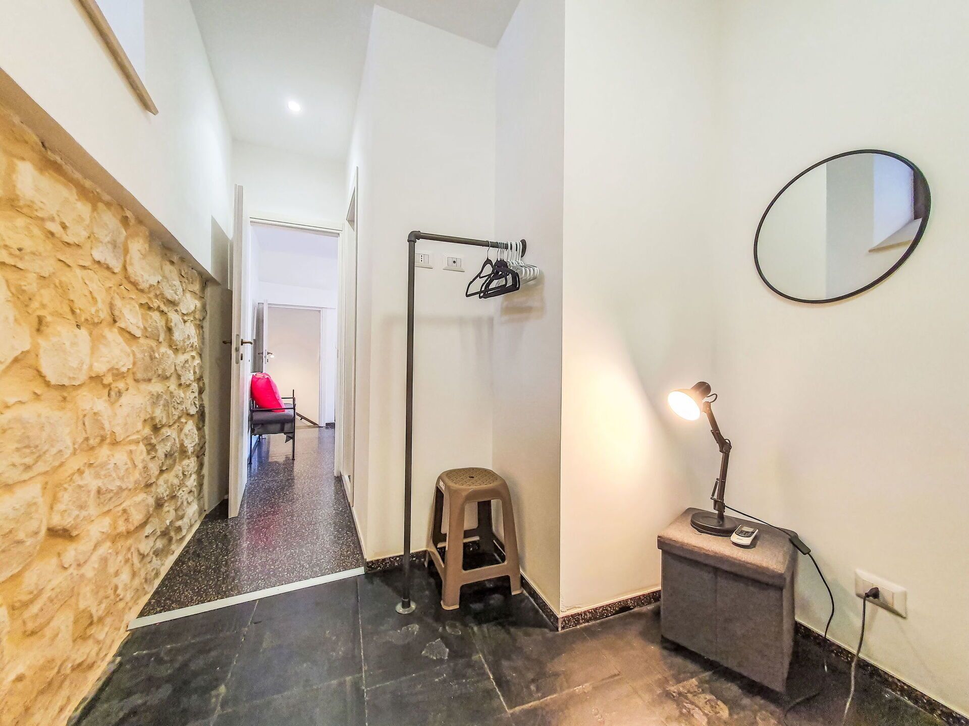 Un couloir avec un mur en pierre, un sol carrelé de noir, un tabouret, un bureau avec une lampe et un miroir accroché au mur.