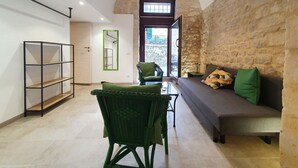 Un amplio salón con pared de piedra, un sofá, un sillón verde, una mesa de cristal y una estantería.