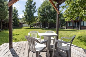 Une terrasse en bois avec une table ronde et quatre chaises, entourée d’une pelouse verdoyante et d’arbres.
