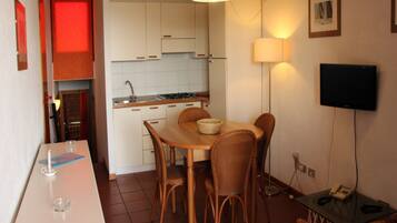 Ett kompakt kök med vita skåp, ett matbord med flätade stolar, en liten köksö och en platt-TV som är monterad på väggen.