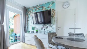 Un comedor moderno con una mesa redonda, sillas acolchadas, un televisor de pantalla plana montado en una pared con estampado floral y vistas al balcón.