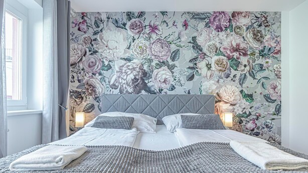 Una cama bien hecha con cabecera gris y papel tapiz con motivos florales, dos lámparas de noche y una ventana con cortinas.