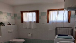 Ein Badezimmer mit Toilette, Bidet und Badewanne. Zwei Fenster mit Vorhängen, ein Handtuchhalter und eine Pflanze an der Wand sind vorhanden.