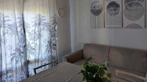 Ein Wohnzimmer mit einem beigefarbenen Sofa, einem Bett mit weißer Bettdecke und einem Fenster mit Palmenvorhängen. Darüber befinden sich drei Wandbilder.