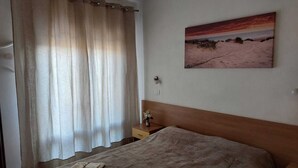 Ein Schlafzimmer mit Bett, Nachttisch, einem Bild mit Strandmotiv und einem Fenster mit Vorhängen.