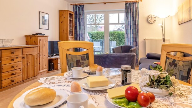 Un comedor con una mesa dispuesta para el desayuno, incluyendo huevos, pan y mantequilla. Hay sillas, un armario y vistas al exterior a través de una ventana.