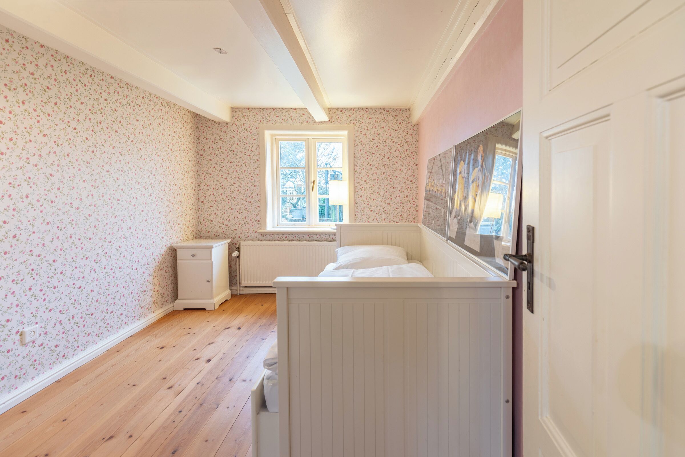 A bedroom with a wooden floor, a window with a view, a radiator, and a white bed.